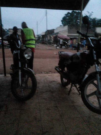 The scene, set in East Ugenya ward, Kenya, depicts an outdoor area at dusk or dawn, characterized by dim, overcast lighting. In the immediate foreground, two motorcycles are partially visible on a dark, possibly paved surface beneath an overhead structure. The motorcycle on the left is largely silhouetted, presenting its front wheel and headlight. Standing beside this motorcycle are two individuals: one is a dark silhouette, and the other, viewed from behind, wears a bright yellow-green reflective vest over a red shirt. The vest exhibits faint text or a logo.
Beyond the foreground, a reddish-brown dirt road extends into the midground, flanked by numerous single-story commercial structures, likely shops or market stalls, with various awnings, including a notable maroon tarp on the right. Several indistinct human figures are distributed along this road, appearing engaged in various activities or loitering. Utility poles are present, suggesting existing infrastructure. The background reveals additional low-rise buildings and scattered trees against a pale, overcast sky.
