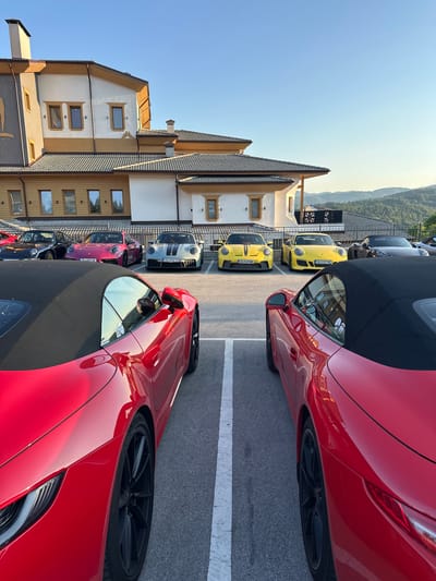 This media file depicts a line of luxury sports cars parked in an outdoor lot. The cars, primarily Porsches, are of various colors, including red, yellow, pink, and silver, and some are convertibles with their tops down. They are parked in front of a building with a distinctive architectural style, featuring tiled roofs, wooden accents, and multiple windows. The building appears to be a hotel or resort, with mountains visible in the background.

The scene suggests a gathering or event involving these vehicles, possibly a car rally or a special occasion. The time of day appears to be late afternoon or early evening, as indicated by the warm, soft lighting and the long shadows cast by the cars and building. The sky is clear and blue, suggesting pleasant weather. The location is identified as Kerenite, Bulgaria.

There are no people prominently visible in the frame, focusing the attention on the vehicles and the surroundings. Notable details include the sleek design of the cars, the pristine condition of their paintwork reflecting the light, and the detailed architecture of the building. License plates are visible on some cars, such as "CB 3127 AE", "CB 2599 WO", "CB 6449 HE", and "CB 2350 TO". The overall atmosphere is one of sophistication and leisure, highlighting the appreciation for high-performance automobiles in a scenic setting.