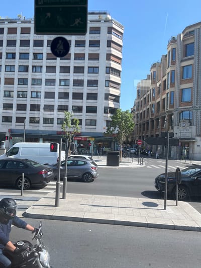 The image depicts a street scene in Neuilly-sur-Seine, France. On the left, a large, light-colored apartment building with numerous windows and brown shutters dominates the background. To the right, a multi-story building with a more ornate, classical facade, featuring brickwork and arched windows, stands. A white van is visible in the middle ground, with traffic lights and pedestrian signals. Several cars are on the road, including a dark sedan, a gray hatchback, and a black sedan. In the foreground, a person wearing a dark blue jacket and a black helmet is riding a motorcycle. The sky is clear blue with a faint contrail from an airplane. The street is paved with asphalt, and there are sidewalks with tiled patterns. Street signs and lampposts are also present. The scene appears to be a sunny day in an urban environment.