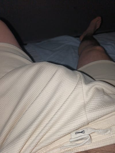 The image shows a close-up view of a person's legs and upper body wearing light-colored, textured shorts. The person is lying down on a bed with blue sheets. The background is dark and out of focus. There is a label with an X-shaped symbol on the shorts. The image does not contain any information about the location in Ümraniye, Turkey.