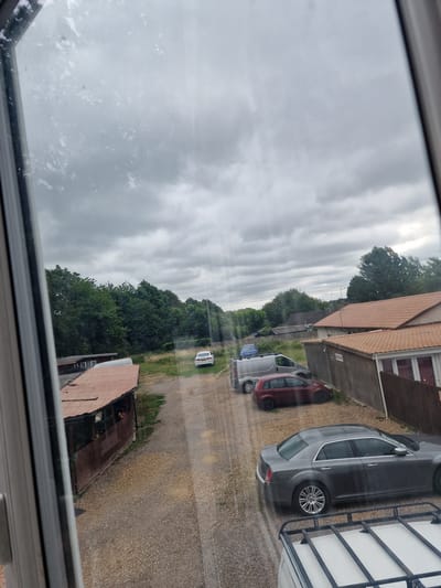 A grey sedan is parked in a gravel lot outside a building with a brown wooden exterior and a red tile roof. In the background, there are trees and a cloudy sky. Several other vehicles, including a silver van and a red hatchback, are also visible in the lot. A long, low structure with a rusty metal roof is situated to the left of the sedan. The scene appears to be in King's Lynn and West Norfolk, United Kingdom.