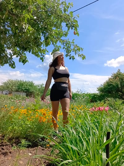 The media file captures a medium-angle shot of a woman standing in a garden under a bright, sunny sky with scattered clouds. She is wearing a white brimmed hat, a black sports bra, black shorts, and a black fanny pack around her waist. Her left hand is slightly extended, and she is looking off to the right, with a relaxed expression.

The garden is filled with a variety of flowers, predominantly yellow and orange, with some pink lilies visible in the background. Lush green grass and foliage frame the foreground and surround the woman. In the distance, behind the garden, are hints of rural structures, possibly houses and a fence, suggesting a residential or rural setting. A large tree with green leaves partially overhangs the scene from the top left.

The lighting indicates it is daytime, likely late morning or early afternoon, given the strong sunlight. The weather appears to be clear and warm. The overall mood is peaceful and serene, with the woman enjoying the natural surroundings. There is no visible text in the media file. The location context is Vyazovka, Russia.