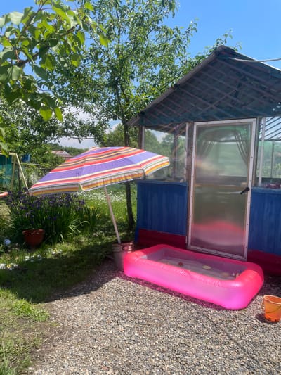 The image depicts a sunny outdoor scene in the city of Yasnoe, Russia, featuring a small, inflatable pink kiddie pool in the foreground. The pool contains a small amount of water and some floating toys. To the left of the pool, a striped beach umbrella is set up, casting a shadow on the gravel ground. Behind the pool and umbrella is a blue and red wooden structure, possibly a shed or playhouse, with a translucent door. The background shows lush green trees and plants, with a bright blue sky overhead. A small orange bucket is visible to the right of the pool.