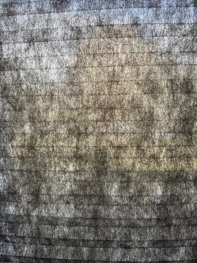 This is a close-up, abstract image of a dark grey, horizontally pleated fabric or material. The pleats are clearly visible, creating horizontal bands of light and shadow. The texture is somewhat rough and fibrous. There are subtle shifts in color, with hints of light grey, white, and a faint yellowish-brown tone in the central area, possibly due to lighting or the material itself. The overall impression is one of texture and pattern rather than a discernible scene. This image is not clearly identifiable as being from Arzamas, Russia, or any specific urban location.