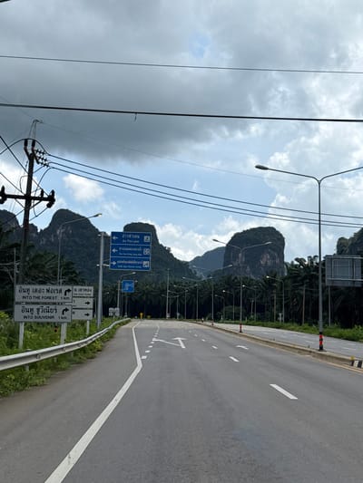 A straight, paved two-lane road stretches into the distance under a cloudy, grey sky. The road is flanked by palm trees and dense green vegetation on both sides. In the background, imposing, dark green karst mountains rise dramatically, their peaks partially obscured by clouds. Several streetlights are visible along the roadside. Traffic signs are mounted on poles, with one set displaying directions in both Thai and English, indicating "Ao Tha Lane," "Tha Pom Klong Song Nam," and "Krabi Agro Tourism Center." Another sign reads "Into The Forest 1 km." and "Into Souvenir 1 km." Overhead, power lines crisscross the sky, supported by a wooden utility pole with insulators. The scene appears to be on the outskirts of Ban Nai Sa, Thailand, characterized by its natural landscape and road infrastructure. There are no people or vehicles visible in the frame.