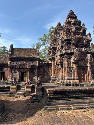 This is a photograph of the Banteay Srei temple in Siem Reap, Cambodia. The temple is made of red sandstone and features intricate carvings and sculptures. In the foreground, there are stone statues of human-like figures. Behind the statues, a staircase leads up to a doorway. The temple architecture is characterized by multiple towers with tiered roofs and decorative elements. The sky is blue with some clouds, and trees are visible in the background. The overall scene conveys a sense of ancient history and detailed craftsmanship.