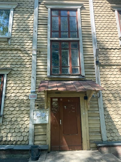 The image shows a two-story building in Novouralsk, Russia. The building has a distinctive facade, composed of hexagonal wooden shingles on the first story and vertical wooden planks on the second story. A window with a brown frame is visible on the left. A brown door is to the right with a piece of rusty metal serving as an awning above the door. Below the door is an aging bulletin board with tattered notices. A sign reading "ОСТОРОЖНО! СХОД СНЕГА!" (Caution! Snow Falling!) is posted next to the bulletin board. A bucket sits next to the building at the base.