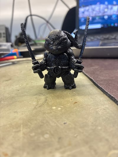 A black 3D printed figurine of a character resembling Raphael from Teenage Mutant Ninja Turtles stands on a textured, light-colored surface. The figurine is holding two swords, one in each hand, and appears to be in a combat stance. The character has a round head with visible texture lines, prominent eyes, and a muscular body. The swords have a detailed, ridged edge. In the background, a laptop screen displays colorful graphics, and other electronic equipment and cables are visible. The overall environment suggests a workshop or laboratory setting. The image does not provide any information about the city of Toruń, Poland.