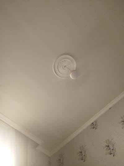 The image shows a white ceiling with a decorative medallion and a small, white, spherical light fixture attached to it. The ceiling is slightly textured. In the lower right portion of the image, a portion of a wall covered in floral wallpaper is visible, meeting the ceiling with white crown molding. The lighting suggests an indoor setting, likely during daylight hours given the bright light coming from the left side of the frame, which appears to be a window. This scene is likely within a residential or commercial building in Sabunchu, Azerbaijan. There are no people or visible actions in the frame.