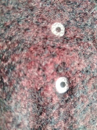 The scene depicts a close-up, static view of two small, metallic objects resting on a textured, multicolored surface. The location is specified as Keekonyokie ward, Kenya.
**Objects:**
Two silver-colored, ring-shaped objects, consistent with beads or spacers, are visible. Each object features a central circular perforation. The outer circumference of both objects displays a textured, possibly hammered or faceted pattern.
1. The upper object is positioned towards the upper-right quadrant of the frame.
2. The lower object is situated towards the lower-center-right quadrant of the frame, approximately directly below the upper object, maintaining a vertical alignment.
**Environment:**
The objects are situated on a fibrous surface, likely textile such as carpet or upholstery. This surface exhibits a mottled coloration, primarily consisting of dark reddish-brown, black, and dark grey hues. The texture appears coarse or shaggy. The background is out of focus, indicating a shallow depth of field.
**People/Actions/Interactions:**
No people, animals, or discernible actions or interactions are present or implied within the frame.
