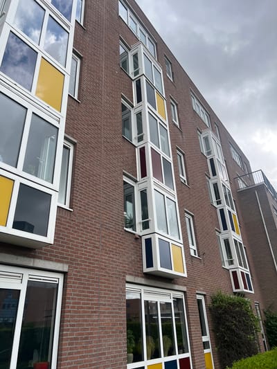 The image shows a close-up, upward-looking view of a brick building with multiple windows, characteristic of modern architecture. The building facade is constructed from dark red bricks, laid in a stretcher bond pattern. The windows are a prominent feature, with many of them being large, rectangular, and framed in white. Some windows are open, allowing a view into the interior spaces, which appear to be apartments or offices. A distinctive detail is the use of colored glass panes in some of the windows, specifically yellow, dark blue, and maroon. These colored sections are strategically placed within the white frames, adding a touch of vibrancy and visual interest to the facade.
The perspective is from below, looking up towards the sky, which is overcast and gray, suggesting a cloudy day. The building is tall, with several stories visible. There are also hints of greenery, with some bushes and possibly ivy growing on or near the building. One section of the building has a more industrial feel with a visible metal structure, possibly a balcony or fire escape, attached to the upper floors.
The scene is likely residential or commercial, given the nature of the windows. There are no people visible in the image. The overall impression is one of urban architecture with a playful and colorful design element. The location context is Leeuwarden, Netherlands. No visible text is present in the image.