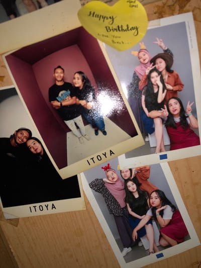 The image displays a flat lay arrangement of five instant photographs and a yellow heart-shaped note on a light brown wooden surface in Tlogo, Indonesia.
A yellow heart-shaped sticky note, slightly curled at the edges, is positioned at the top center. It bears handwritten text: "Happy Birthday Love You Best..." with smaller text "alfad amel" above.
The photographs include:
1. **Top-left:** Partially visible, out-of-focus portion of an individual.
2. **Mid-left:** A portrait-oriented photo featuring a male and a female, both smiling at the camera, with their heads close together against a dark background. The male wears a dark top; the female wears a dark top. The word "ITOYA" is printed at the bottom of the white border.
3. **Center:** A larger portrait-oriented photo shows a male and female standing inside a small, maroon-walled room with a white floor. The male, on the left, wears a black t-shirt, splatter-painted light pants, and light shoes, with arms crossed and a slight smile. The female, on the right, wears a dark long-sleeved top and blue jeans, with her arm extended towards the male, also smiling. A bright light reflection obscures the lower right portion of the photo. The word "ITOYA" is printed at the bottom of the white border.
4. **Top-right:** A portrait-oriented photo of five young women posing against a light gray background. One woman wears a pink hijab with orange bunny ears and makes a peace sign. Another wears a brown hat and a brown shirt, also making a peace sign. A third woman sits, wearing a dark top and blue skirt, with one hand near her face. A fourth woman in a dark red dress makes a peace sign with her tongue slightly out. A fifth woman's face is partially visible in the background.
5. **Bottom-right:** A portrait-oriented photo of five young women posing against a light gray background. One woman, wearing a pink hijab with red crab-claw headwear and a patterned dress, leans her head against her hand with a fish-face expression. Another woman in a brown shirt is partially visible. A third woman, with long dark hair, wears a dark top and blue skirt, sitting. A fourth woman in a dark red dress with a white collar makes a pouty face. A fifth woman's face is partially visible. Blue horizontal stripes are printed at the bottom of the white border.