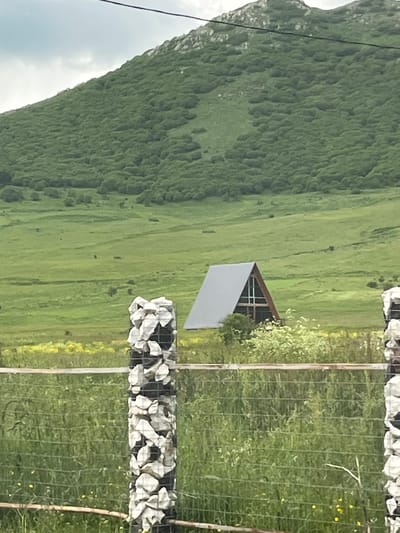 An A-frame house with a gray roof is nestled in a lush green meadow at the base of a tree-covered mountain. In the foreground, a wire fence runs across the image, with two decorative posts made of stacked white stones. The sky is overcast, and the overall scene depicts a rural landscape in Lusagyugh, Armenia.
