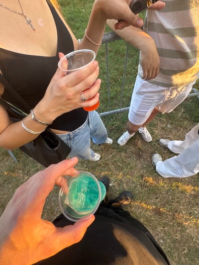 The overhead shot shows a group of people gathered outdoors on a grassy area, possibly at a festival or gathering. The time of day appears to be late afternoon or early evening, indicated by the warm, golden sunlight casting long shadows.
The foreground features two individuals. One person, wearing a black top and jeans, is holding a clear plastic cup filled with a blue-green beverage and ice cubes. They are also wearing a silver cross necklace and several silver bracelets. Another person, with visible arms and hands, is holding a clear plastic cup with an orange-colored liquid, and is wearing a ring on their finger and a pink bracelet.
Further in the background, two other individuals are visible. One person is wearing white pants and white sneakers. Another person is wearing a striped blanket or towel over their legs and white shorts, also with white sneakers. The legs and feet of another person wearing black pants and dark shoes are also visible.
The scene suggests a casual, social event. The grass and outdoor setting imply an informal atmosphere. The presence of drinks and the relaxed postures of the individuals indicate a relaxed and possibly festive occasion. No visible text is present in the image.