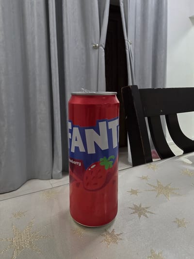 A red Fanta can with a strawberry graphic and "Fanta Strawberry" text is placed on a white tablecloth with gold star patterns. The can is the primary subject of the image. In the background, gray curtains hang. Part of a dark wooden chair is visible on the right side of the image. The scene appears to be indoors, possibly in a dining area or living room. There are no people present. The image does not provide any specific information about the location being in Dibba Province, Oman.
