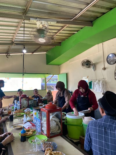 The image depicts a bustling interior scene at a restaurant in Margoyoso, Indonesia. The dining area is open-air with a corrugated metal roof supported by metal beams, painted green. Two large ceiling fans are visible, suggesting a warm climate. The walls are painted cream, with green accents. On the right wall, a round analog clock with black numbers and hands is prominently displayed.
The central focus of the image is a long table laden with food and drinks, where patrons are seated and served. Several people are actively involved in the dining and serving process. Two women, wearing headscarves and black aprons, appear to be serving food. One is scooping rice from a large green container into a plate held by a man. The other woman is tending to food preparation behind the counter.
On the table, various dishes are arranged, including plates of fried chicken, crackers, and bowls of soup. Bottles of water and drinks are also visible, along with condiments. The patrons, a mix of men and women, are engaged in eating and conversation. One man, wearing a black and white checkered shirt and a peci (a brimless cap), is facing away from the camera, interacting with the servers.
The overall atmosphere appears to be that of a casual, local eatery, likely a popular spot for meals in Margoyoso. The scene captures a moment of community and dining, typical of Indonesian culinary culture.