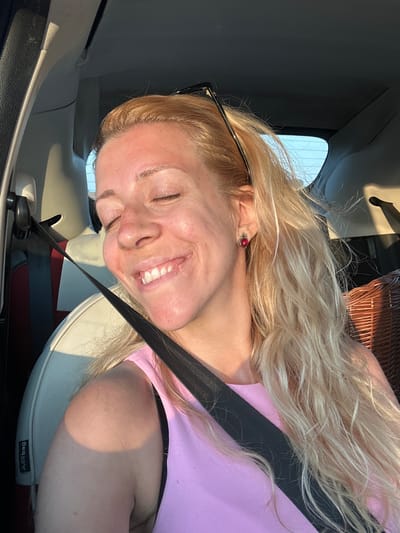A blonde woman is sitting in the passenger seat of a car. She is wearing a pink top and a seatbelt. Her eyes are closed and she is smiling. The sun is shining through the window, casting a shadow on her arm. The car is likely on a road in Alvimare, France, as indicated by the presence of a wicker basket, commonly used for picnics and outings in rural French settings.
