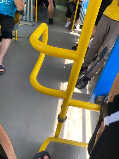This media file captures an interior view of a bus, possibly in Zvezditsa, Bulgaria. The scene is dominated by a bright yellow handrail, a prominent feature for passengers to hold onto. Several people are visible, though their faces are not the primary focus. One person is wearing a yellow t-shirt and grey trousers, and another is in a blue t-shirt and black shorts. Legs and feet of other individuals are also present, indicating that the bus is in motion or stopped with passengers boarding or disembarking.

The lighting suggests it is daytime, with sunlight casting shadows and creating bright patches on the floor, hinting at a sunny day. The setting is clearly a public transportation vehicle, indicated by the seating, the handrail, and the general interior design. There is a blue seat or panel visible in the upper portion of the image.

One visible detail is text on the blue t-shirt, which appears to be in a foreign language, possibly Bulgarian given the location context. The overall impression is a candid snapshot of everyday life on a bus. No specific activity or event is clearly defined, but the presence of passengers suggests typical travel. The scene is mundane, without overt displays of emotion. The focus is on the functional elements of the bus interior and the presence of people within that space.