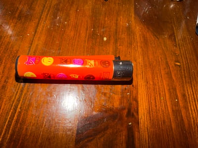 An orange Clipper lighter with emoji and cartoon graphics sits on a stained wooden surface. The background is a medium-toned, polished wood with varied grain patterns. It is impossible to determine from this image if the location is in Khalde, Lebanon.