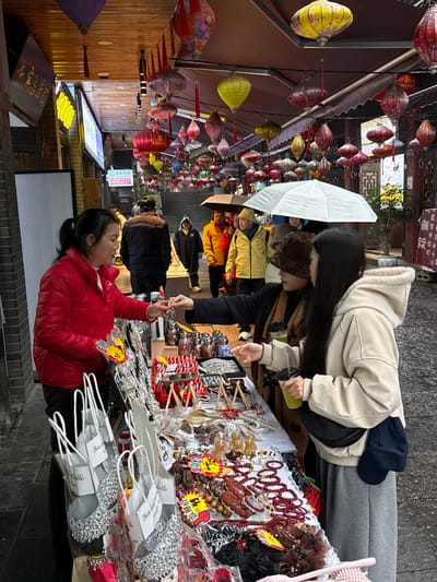 The image depicts an active outdoor market scene on a wet, paved street or alleyway within Pengshui Miao and Tujia Autonomous County, China. The overhead area is extensively adorned with numerous colorful traditional Chinese lanterns of varying shapes and sizes, suspended from wires and architectural elements.
In the foreground, a female vendor, appearing middle-aged and wearing a bright red puffer jacket, stands behind a white-topped stall displaying a diverse range of merchandise. She is engaged in an interaction with two younger women, presenting or handing a small, silver-colored item to one of them. The stall items include hanging silver-colored ornaments, small containers with colorful contents, red-and-white striped objects, wooden spoons, carved figurines, multiple beaded bracelets (predominantly red and brown hues), and various hair accessories. Several white paper bags containing silver-colored items are visible to the vendor's left.
The two female customers stand to the right of the stall. One woman, with long dark hair, wears a light beige hooded jacket, grey sweatpants, and a black fanny pack, holding a clear cup with a green beverage. The other woman, partially obscured, wears a dark coat, a brown furry hat, and also holds a clear cup with a green beverage.
In the background, the street inclines uphill, transitioning into stone steps. Multiple pedestrians are visible, some walking with open umbrellas (one prominent white umbrella, and several darker ones), indicating wet conditions, likely from recent or ongoing rain. Individuals in the background are dressed in various casual outerwear, including a bright yellow jacket and dark puffer jackets. Buildings with traditional Chinese architectural features, including wooden elements, line both sides of the thoroughfare. An illuminated sign with yellow characters is visible on a building to the left, and a traditional wooden doorway and foliage are seen to the right.