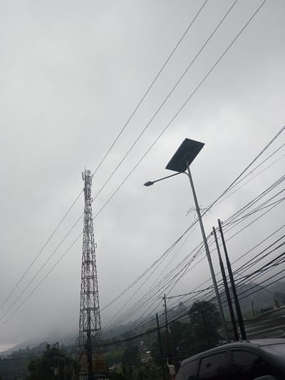 A tall, metal communication tower stands prominently against a cloudy sky. Electrical wires crisscross the scene, adding to the dense network of infrastructure. A solar-powered street light illuminates the right side of the image. In the background, a lush hillside is partially obscured by fog, creating a sense of depth. The overall atmosphere is overcast and industrial.