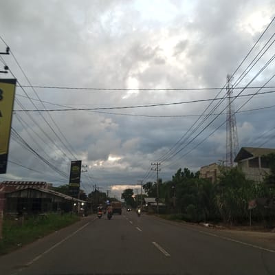 This image depicts a view looking down a two-lane asphalt road in Payameuneng, Indonesia, under a heavy, overcast sky, likely at dusk or dawn. The sky is dominated by thick, grey clouds, with brighter patches indicating obscured sunlight towards the upper center and right.
On the left side of the road, in the foreground, is a dilapidated single-story wooden building with a rusty corrugated metal roof, partially obscured by power poles and a large, vertical yellow billboard. Green grass and foliage grow along the roadside. Further back on the left, more structures and dense overhead utility wires are visible.
The right side of the road features abundant green vegetation, including trees and tall grasses, obscuring views of buildings behind them. A prominent, dark vertical billboard with some text and "19.000" is visible in the mid-ground, positioned near power poles. In the background on the right, a tall, lattice-structured telecommunications tower extends high into the cloudy sky. Buildings are partially discernible behind the foliage and in the distance.
The road itself has a solid white line on its left edge and dashed white lines marking the center, indicating two lanes of traffic. Several vehicles are in motion: a light-colored truck is visible in the distance on the left lane, moving away from the viewer. Multiple motorcycles are on the road; at least one motorcycle with a rider and illuminated headlight is in the foreground on the left lane, moving away. On the right lane, several motorcycles are visible, some with illuminated headlights, approaching the viewer. The overall lighting is dim, suggesting low light conditions, with artificial lights from buildings and vehicle headlights active. A dense network of black utility wires crisscrosses the upper half of the image between numerous power poles.