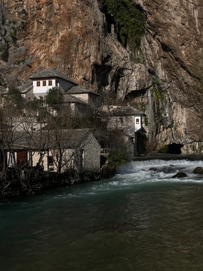 The image displays the Blagaj Tekke complex at the spring of the Buna River in Dračevice, Bosnia and Herzegovina. A formidable, multi-textured rock cliff, predominantly brown and grey, forms the background, featuring scattered green vegetation and a prominent dark cave entrance at its base on the right. Below the cliff, a cluster of traditional stone-built structures with grey slate roofs are integrated into the terrain. A two-story building with white plastered walls and three brown-framed windows on its upper level is centrally positioned. To its right, another white-walled building stands directly at the cliff's base, adjacent to the river. The Buna River, characterized by its clear turquoise water, flows from right to left in the foreground and mid-ground. A distinct waterfall or weir with white rapids is visible in the mid-ground, creating turbulent water. Along the left bank of the river, a stone building features a covered outdoor patio area equipped with tables and chairs. Numerous bare deciduous trees are interspersed among the buildings and along the riverbanks, indicating a seasonal lack of foliage. No individuals are present.
