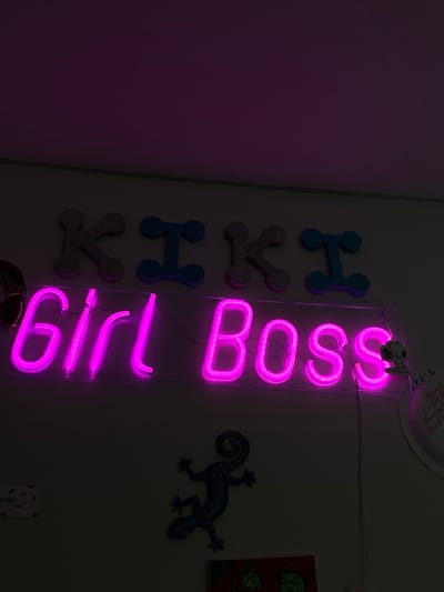 The image depicts a dimly lit interior wall within a private residence in Ridgefield, United States. Mounted on the light-colored wall, an illuminated pink neon sign spells "Girl Boss" in script font, affixed to a clear rectangular backing. Above this sign, individual decorative letters spell "KIKI"; the 'K's are textured and glittery, while the 'I's are solid blue with rounded ends. Below and to the left of the neon sign, a blue-green, patterned lizard decoration is affixed to the wall. Further down, partially visible on the lower wall, are a small white rectangular object with red heart patterns on the left, and a circular white paper note or drawing on the right with visible text "Most likely to...". Below the lizard, a dark, rectangular piece of art featuring green figures is partially visible. No individuals or specific actions are present.