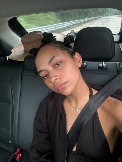 A female individual with medium skin tone and dark hair styled in two buns is depicted seated in the rear passenger compartment of a vehicle. She wears a black halter-neck top, an open black hooded jacket, a silver-colored necklace with star pendants, and a septum piercing. A black seatbelt diagonally crosses her torso. Her expression is neutral as she looks directly at the camera. The vehicle interior features black seats and headrests. A light brown straw hat rests on the rear parcel shelf. Through the rear window, a road with a metal guardrail and green foliage is visible, indicating a rural or highway environment under overcast conditions. The image provides no discernible details to confirm the specific location as Colonia Guerrero, Mexico.