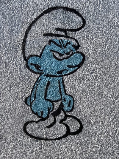 The scene, located in Tomé, Chile, displays a piece of graffiti art on a textured, light gray stucco wall. The central subject is a full-body depiction of the cartoon character, Grumpy Smurf. The character is rendered in light blue with a prominent black outline, while its characteristic hat and pants are uncolored, appearing white against the wall's base tone. Grumpy Smurf is depicted with a scowling expression, featuring furrowed brows, narrowed eyes, and a downturned mouth. Its arms are bent at the elbow, and hands are positioned at waist level. The wall exhibits a coarse, irregular texture, illuminated by direct, bright lighting.