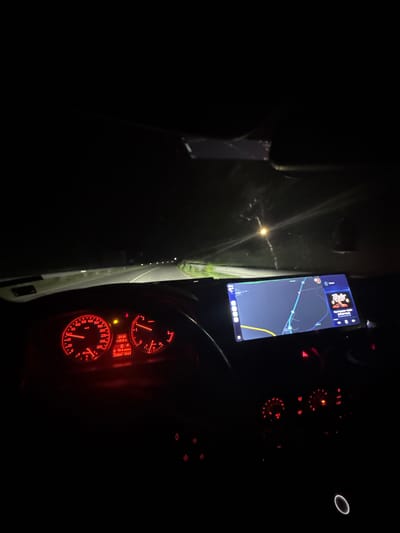 Captured from inside a vehicle at night, the scene focuses on the car's interior dashboard and a navigation screen affixed to the windshield. The dashboard glows red, displaying the speedometer, tachometer, and other essential indicators, showing "23:34," a temperature of "19.5 C," "D" gear, "10.4 km," and an odometer reading of "329051 414.5". A phone-sized navigation screen is mounted on the windshield, displaying a map. Outside the vehicle, a dark road stretches into the distance, illuminated by dim streetlights.