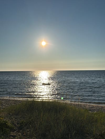 The image shows a serene seascape at Meremõisa, Estonia, under a clear blue sky. The sun is positioned high in the sky, casting a brilliant reflection on the water's surface, creating a shimmering path of light. A small boat with two people can be seen in the distance, navigating the gentle waves. The foreground features a sandy beach with tall grasses, adding a natural and tranquil element to the scene. The overall atmosphere is peaceful and idyllic, capturing the beauty of a summer day by the sea.