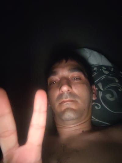The image captures an adult male in a dimly lit, indoor setting, likely a bedroom, located in Servas, France. The man is seen from the chest up, reclining, and appears shirtless, displaying chest hair. He has dark, short hair, prominent eyebrows, and stubble on his chin and upper lip. His eyes are open and directed towards the camera. His left hand is raised in the immediate foreground, forming a "V" shape with the index and middle fingers. A pillow with a dark, swirled pattern is visible behind his head to the right of the frame.