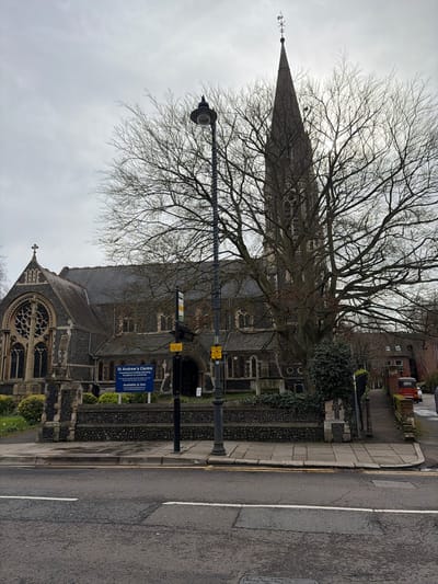 This image depicts St Andrew's Church in Hertford, East Hertfordshire, United Kingdom, under an overcast sky. The prominent Gothic Revival church, constructed of dark grey masonry, features a tall, slender spire topped with a weather vane and a large rose window on its visible west facade. A substantial deciduous tree with bare branches stands in the midground, partially obscuring the church's central tower and upper sections.
In the immediate foreground, an asphalt road with white lane markings abuts a paved sidewalk, which is edged by a yellow curb line. A black lamppost with a circular lantern fixture is centrally positioned on the sidewalk. Behind it, a low stone wall forms the perimeter of the church grounds. A blue rectangular sign within the grounds identifies the building as 'St Andrew's Centre,' promoting 'wellbeing' and noting its availability 'to hire.' Several yellow warning or informational signs are affixed to the lamppost structure. No individuals are present in the scene. To the far right, a portion of a red vehicle and residential architecture are visible.