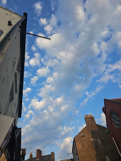 Looking up at the sky with buildings on either side. The sky is blue with white clouds. A wire stretches across the sky from left to right. On the left, there is a white building with a black roof, a black pole sticking up from the roof, and a black rain gutter. On the right, there are several brick buildings, one with a round window. The photograph appears to be taken in Holywell, United Kingdom, given the buildings and the location tag.