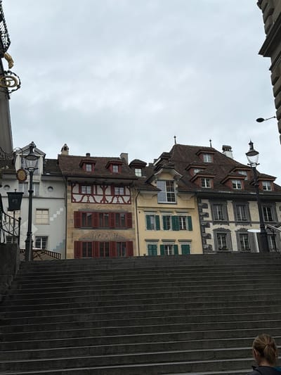 The image is an angled, vertical view of buildings in Lucerne, Switzerland. The architecture features a combination of timber-framed and stucco facades, with varying window designs and shutter colors. Several buildings have pitched, brown tile roofs. A portion of a concrete structure with vertical grooves is visible to the right, obscuring part of the buildings. In the lower portion of the image, there's a glimpse of ornate wrought ironwork and a hanging sign. The sky is overcast. Part of a person's head is visible in the upper right corner.