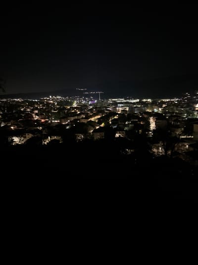This is a nighttime shot of a coastal city, potentially Sveti Konstantin, Bulgaria. The image is taken from a high vantage point, possibly a hill or elevated ground, looking towards the coast. The cityscape is illuminated by artificial lights from buildings, streets, and possibly harbor installations. The dark areas suggest wooded areas or undeveloped land between the viewpoint and the lit-up urban landscape. The sky is dark, devoid of significant lighting. There is no indication of human presence in the immediate foreground. The scene lacks people, subjects, objects, actions, or interactions. The essential elements are the dark environment, the lighted city, and the elevated viewing angle.