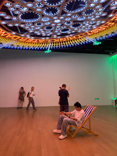 In a brightly lit indoor space, possibly an art gallery, in Jinniu District, China, a circular art installation hangs from the ceiling, adorned with numerous white lights arranged in circular patterns and outlined by yellow and blue neon lights. Below, the polished wooden floor reflects the ambient light.
Four individuals are present. In the foreground, a young man with black hair is seated in a striped deck chair, wearing a white t-shirt, blue jeans, and white sneakers, and appears to be looking at his phone. Behind him, to the left, a man in a dark blue t-shirt and shorts stands, holding a phone and a camera, seemingly taking a picture or recording. Further left, two young women stand against a white wall. The woman on the left wears a patterned dress and sandals, with her hand on her hip. The woman on the right wears a white t-shirt, grey pants, and sandals, carrying a backpack and holding a phone.
The environment is minimalist with a large white wall dominating the background. A green light emanates from the right side of the space, creating a vibrant glow. An exit sign is visible on the wall to the right. The overall impression is one of a modern exhibition space.