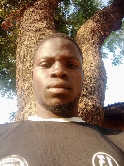 This image captures a close-up portrait of an adult male in an outdoor setting within Yammata, Nigeria. The individual, positioned centrally, has a dark skin tone, short dark hair, dark eyes, a prominent nose, full lips, and a short, dark beard and stubble outlining his chin and jawline. He wears a black short-sleeved shirt featuring a white collar, with a white circular logo partially visible on the lower right quadrant of the shirt. His expression is neutral as he looks directly into the camera.
The background is dominated by the textured, dark bark of a large tree trunk, which splits into two substantial branches directly behind the subject. Green foliage is present on the tree branches, particularly on the left and upper right sides of the frame. A piece of patterned fabric, white with brown and orange designs, is draped over a branch in the upper left background. A bright, clear sky is visible through the gaps in the tree's canopy. A dark structure, possibly a corrugated roof, is faintly visible in the far right background, beneath the tree's foliage. The scene is illuminated by natural daylight.
