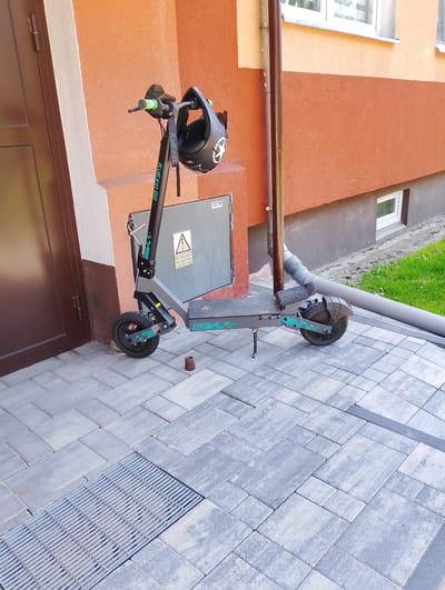 An electric scooter is parked on a paved walkway next to a building with an orange facade. A black helmet with a white logo is hanging from the scooter's handlebars. A downspout runs vertically along the building. A metal grate is visible in the foreground. The scene is set in the city of Kolno, Poland.