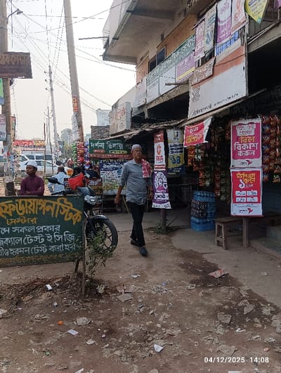 This image depicts a bustling street scene in Kaliganj, Bangladesh, recorded at 14:08 on December 4, 2025. The immediate foreground shows uneven, compacted dirt ground extensively littered with paper fragments, plastic, and small debris. On the left, a vertical green sign with white Bengali text, likely indicating "F. Diagnostics" and listing medical tests, stands next to a small green plant rooted in the dirt.
A male subject, appearing middle-aged to elderly, walks forward in the center of the frame. He wears a light grey short-sleeved collared shirt, dark trousers, dark shoes, and a white skullcap, with a red and white checkered cloth draped over his left shoulder. To his left, another male subject, appearing younger, is seated on a low wooden bench, dressed in a maroon long-sleeved shirt and a white skullcap. A dark-colored motorcycle is partially visible behind the seated individual.
The midground features a commercial establishment on the right, displaying numerous bags of snacks hanging from an awning. Multiple colorful banners and posters, primarily in Bengali, are affixed to the building and utility poles, advertising services such as "Bikash" and various products. A stack of blue plastic water bottles is visible near the store's entrance, adjacent to a small, dark wooden stool.
The street extends into the background, lined with numerous utility poles supporting a dense network of overhead electrical and communication wires. Further down the street, additional indistinct figures, a white van, and other vehicles are present. The background also includes a multi-story concrete building with an overhanging roof structure. The sky overhead appears bright and hazy.