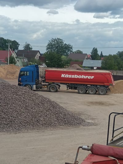 The image depicts a gravel pit or construction material storage site in Яльгелево, Russia. A blue semi-truck with a red trailer bearing the name "Kässbohrer" is the central subject, with the trailer filled with sand or gravel. A person is standing by the truck cab. There are large piles of gravel and sand visible in the foreground and background. The backdrop shows residential houses and trees under a cloudy sky.