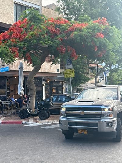 The image depicts a street scene in Kfar Shmaryahu, Israel, dominated by a large tree with vibrant red flowers and green foliage. In the foreground, a silver Chevrolet pickup truck is parked. To the left, a section of a building with outdoor seating for a cafe is visible, with several individuals seated at tables. A white umbrella is partially unfurled near the entrance. A compact, four-wheeled electric vehicle is parked beside the tree. A yellow sign with Hebrew text is mounted on a pole near the truck. Two Israeli flags are visible in the background. The setting appears to be a commercial or public area within the city.