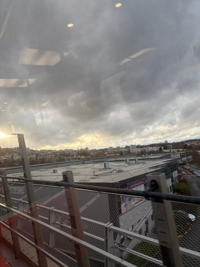 A cloudy sky dominates the scene, partially obscured by window reflections. The cityscape below includes buildings, a parking lot with cars, and distant trees. A metal structure, possibly scaffolding or a railing, is visible in the foreground. Light shines through the clouds, casting a bright glow on the horizon. The image appears to be taken from an elevated position, looking down at the city.