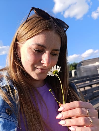 A young woman with long, auburn hair and freckles on her face is shown in a close-up shot, holding a white daisy with a yellow center. She is looking down at the flower with a gentle smile, her eyes closed. She wears a purple t-shirt and a denim jacket. Her fingernails are painted with a pink and white gradient, and she is wearing two rings on her right hand. Sunglasses are perched on top of her head. The background features a bright blue sky with fluffy white clouds, suggesting a sunny day. In the blurred background, there are some dark wooden benches and a silver trailer or vehicle, indicating an outdoor setting, possibly a rural area or park. The lighting suggests it is late afternoon or early evening, with the sun casting a warm glow on her face. The overall mood is peaceful and serene. The location is identified as Ivanovskoe, Russia. There is no visible text in the image.