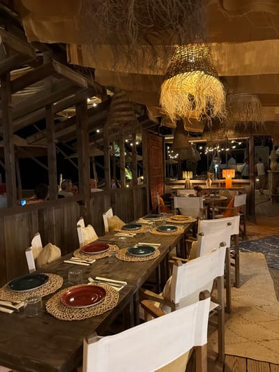 The image depicts the interior of a restaurant in Torreilles, France, characterized by a rustic and natural aesthetic. The scene is illuminated by warm, ambient lighting emanating from large, woven pendant lamps with frayed edges hanging from a beamed ceiling. The ceiling itself is draped with tan fabric, further contributing to the earthy ambiance.
In the foreground, a long, dark wooden table is set for dining. Each place setting includes a woven placemat, a red or dark green plate, silverware, and a glass. Several chairs with white canvas coverings are arranged around the table.
In the background, the restaurant's architecture is visible, featuring wooden beams and partitions. The restaurant appears to be open-air or have large openings, as evidenced by the view of the night sky with string lights and silhouettes of palm trees. Some patrons are visible in the background, seated at tables or standing. One person is holding a smartphone. The overall impression is that of a relaxed, beachside or tropical-themed dining establishment.