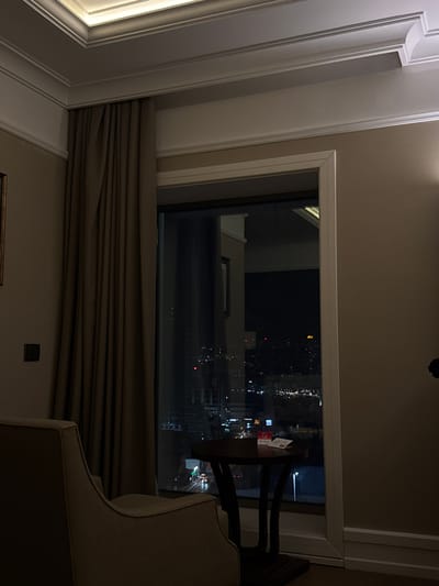 A view from inside a room overlooking a city at night. The room has curtains, a chair, and a table next to the window. The lights of the city are visible through the window. It is not possible to determine whether the image is of Bağcılar, Turkey based on the image content.