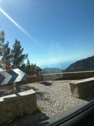 Captured from inside a vehicle, the image shows a roadside view in Fornalutx, Spain. The perspective is from within the vehicle. The road surface is rough and textured. A concrete barrier runs along the edge of the road. A blue and white chevron sign indicates a sharp turn. Lush greenery and a steep mountain face the roadside. The sky is clear and blue.
