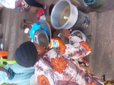 The scene depicts an outdoor food preparation or serving event in Kimamba, Tanzania. In the foreground, a person, identifiable as female, is observed wearing a black head covering and a long-sleeved floral top with orange, brown, and white patterns. This individual is actively scooping food with a silver spoon from a small metal pot, which contains a light-colored solid food, into another, partially obscured, metallic dish.
To the left of the central figure, a stack of serving dishes is visible, consisting of a large, circular, silver-colored metal plate resting atop a blue plastic bowl and other plates. An orange plastic bottle stands nearby. In the background to the right, a large, open, silver metal pot is situated on a dark, cylindrical cooking stand, containing a light-colored liquid with visible solid contents.
Other individuals are present in the immediate vicinity. To the left, a person wearing a white t-shirt with red lettering and patterned bottoms is partially visible. Further left, a third person in light blue attire is seated on a green plastic chair. A dark green plastic bucket is visible in the far background. The ground consists of an uneven surface appearing to be packed earth or worn concrete, exhibiting cracks and debris.