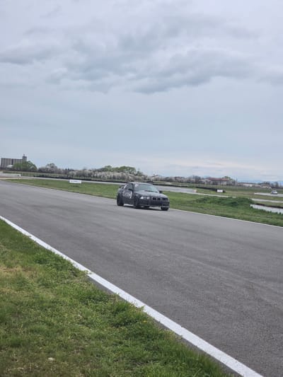 The image shows a black BMW sedan on a racetrack in Kaloyanovo, Bulgaria. The car is on a grey asphalt track with white lane markings on the right edge. Green grass grows alongside the track. In the background, there is a line of trees and buildings under a cloudy sky.